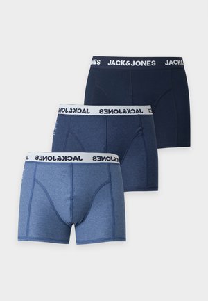 Modèle masculin debout pieds nus en boxer bleu Jack & Jones sur un fond blanc uni, regardant sur le côté.
