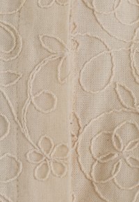 Tissu crème avec broderie florale texturée en motifs bouclés et une couture visible courant verticalement au centre.