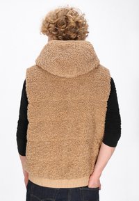 Gilet in pile beige con colletto alto, superficie testurizzata e orlo elastico, abbinato a una maglietta nera a maniche lunghe e jeans blu scuro.