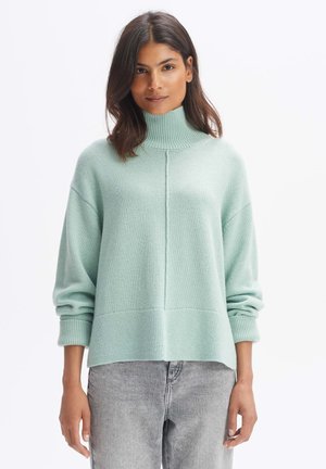 Mintgrüner, gestrickter Rollkragenpullover mit lockerer Passform, gerippten Bündchen und einer Naht, die vorne verläuft. Kombiniert mit grauen Jeans.