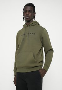 Olivovo zelená mikina s kapucňou, s prednou vreckom a námořnickým logom "TOMMY JEANS". Kapucňa so stiahnuteľnou šnúrkou. Mäkká textúra látky.