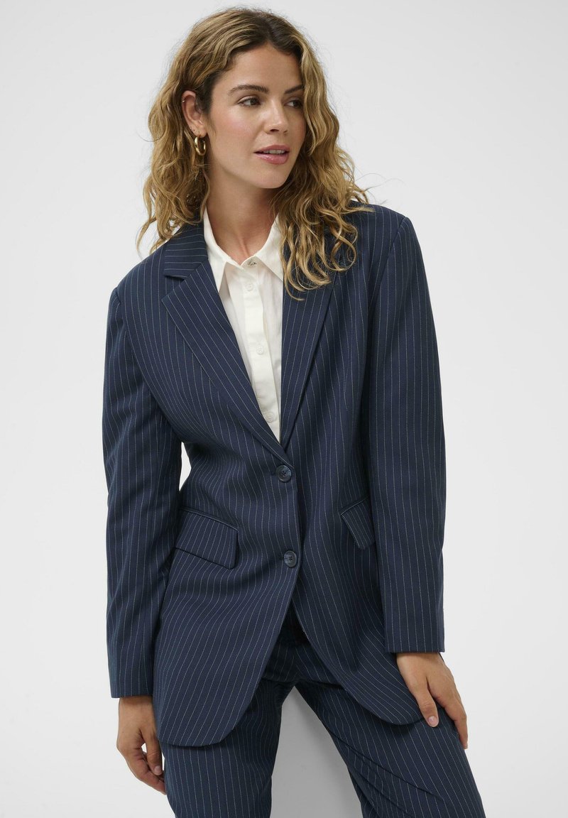 Tengerészkék tűszegélyes blézer két gombbal, bevágott hajtókákkal és két első zsebbel. Sima anyagból készült, testre szabott szabással.