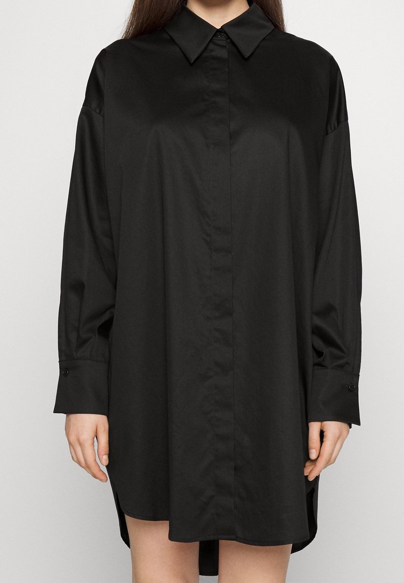 Femme portant une robe-chemise noire oversize à manches longues avec un col pointu et des poignets boutonnés sur un fond uni.