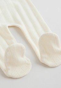 Collants pour bébé en maille côtelée blanche douce avec des couvre-pieds texturés en forme de cœur, posés à plat sur une surface blanche.