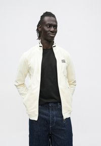 Lys kremfarget jakke med glidelås og opprettstående krage, teksturert stoff og svarte detaljer, kombinert med en svart T-skjorte og mørke jeans.