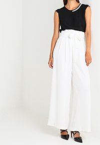 Top sin mangas negro con acento plateado, combinado con pantalones anchos blancos atados en la cintura. Zapatos de tacón alto negros completan el conjunto.