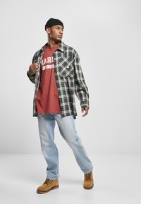 Zeleno in kremasto karirasto flanelasto srajco preplavljena čez temno rdečo majico z grafiko, svetlo modre jeans hlače in svetlo rjave vezalke. Črna kapa.