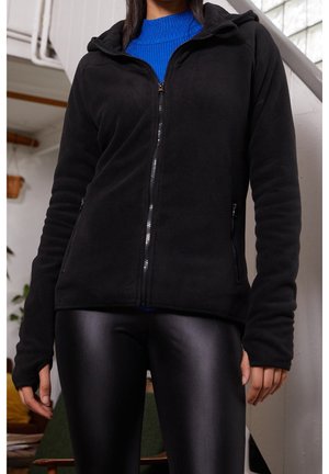 Frau trägt eine schwarze Fleecejacke mit Reißverschluss über einem blauen Pullover und schwarzen Leggings mit Lederoptik, steht drinnen in der Nähe einer Treppe.