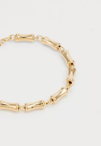 Bracelet doré avec des perles rectangulaires arrondies enchaînées sur un fond blanc.