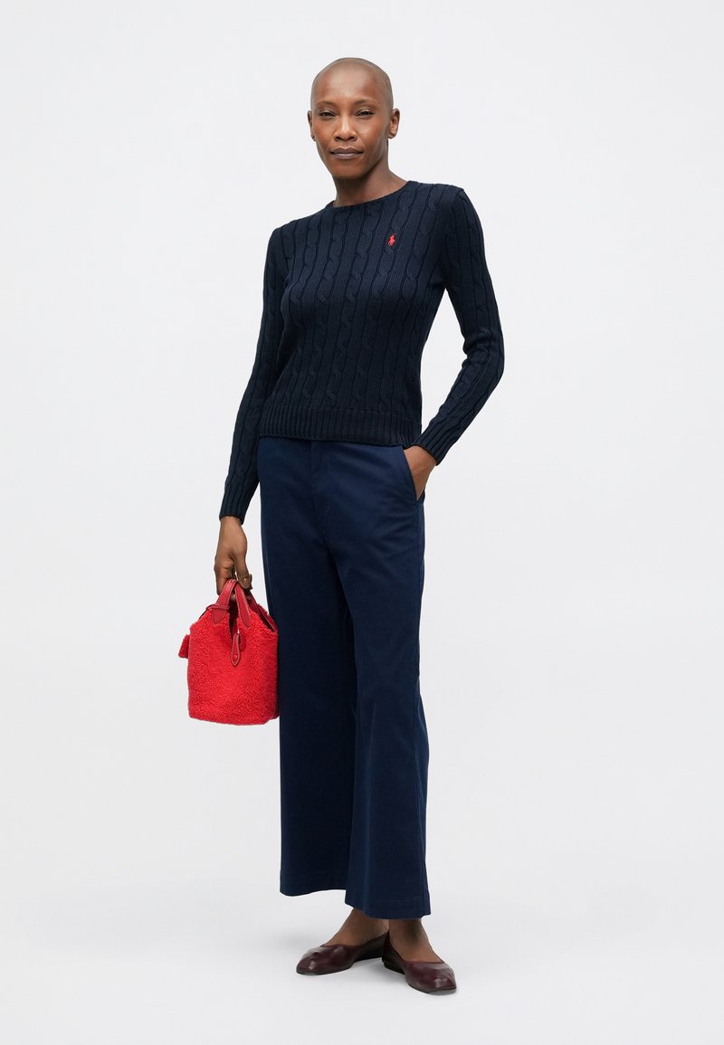 Pull en maille torsadée navy, pantalon navy large, ballerines bordeaux et sac texturé rouge. L'ensemble présente un style ajusté avec des poignets côtelés.