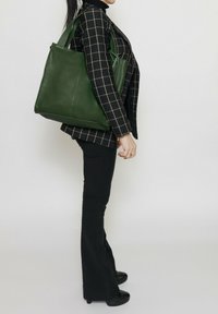 Borsa tote in pelle verde con forma strutturata, doppi manici e texture liscia. Modello mostrato con giacca e pantaloni a quadri neri.