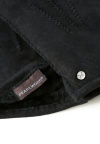 Juodas dirbtinės kailio audinys su minkšta tekstūra, su metaliniu mygtuku ir etikete "PEARLWOOD®." Matomas siuvinėjimo rašto detalės.