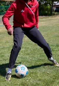 Felpa rossa Adidas con cordini bianchi, pantaloni sportivi neri e scarpe da calcio nere e gialle, posizionata vicino a un pallone da calcio blu e bianco su erba.