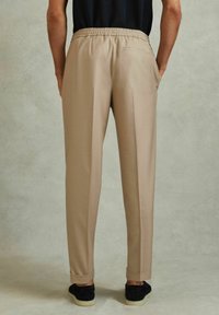 Pantalones beige con cintura elástica y corte ajustado, con puños enrollados y un bolsillo trasero. Combinados con zapatillas de ante negras.