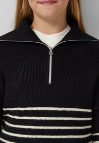 Schwarzer gerippter Pullover mit Reißverschlusskragen, weißem Kragen und horizontalen weißen Streifen über der Brust. Weicher Stoff und taillierte Passform.