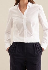 White long-sleeve button-up shirt with a slim fit, featuring an embroidered black rose detail at the lower left. Paired with dark trousers.
