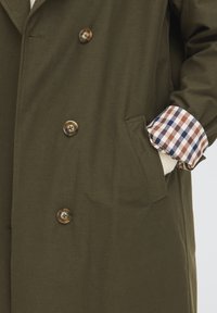 Main dans la poche d'un manteau vert olive avec des boutons marron et une manche retroussée montrant une doublure à carreaux marron, bleu et blanc.