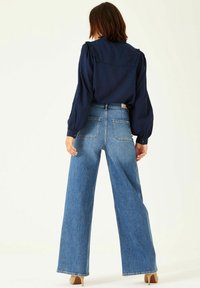 Marinblus med långa ärmar och volanger vid axlarna, tillsammans med höga, vida blå jeans. Slät denimtextur, klassiska fickor.