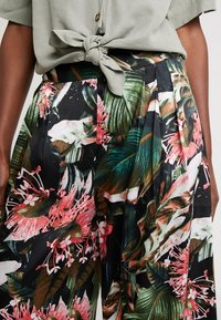 Bloemige plissébroek met levendige groene bladeren en roze bloemen op een zwarte achtergrond, gecombineerd met een vastgebonden saliegroen topje.