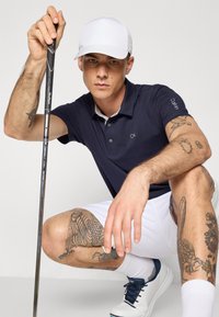 Námornícka polokošela s logom CK, biele šortky a námornícke topánky. Drží golfovú palicu, zobrazuje podrobné tetovania na ramenách a nohách. Nosený biely klobúk.