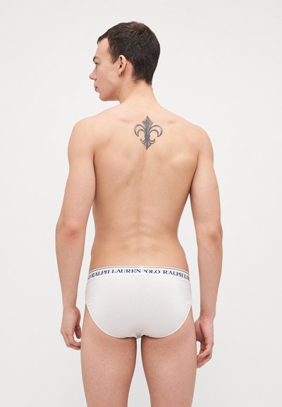 Beli bombažni hlačniki z temno modrim pasom z blagovno znamko "Polo Ralph Lauren". Model ima tetovažo na zgornjem delu hrbta.