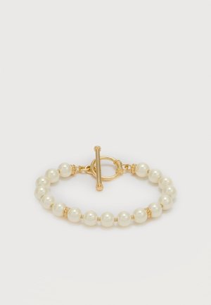 Brățară cu perle albe rotunde și mărgele aurii mici, cu un închizător decorativ din aur pe un fundal simplu și deschis.