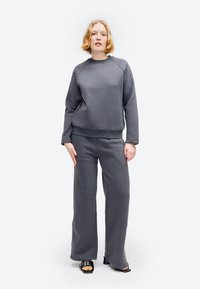 Lyshåret kvinde stående iført mørkegrå langærmet sweatshirt og matchende vidde bukser med sorte åbne hæle på hvid baggrund.