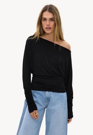 Femme portant un haut noir à manches longues décolleté épaules avec des manches froncées et un jean bleu clair taille haute à jambes larges.