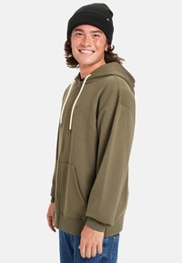 Sudadera con cremallera en color caqui, con capucha ajustable, bolsillo tipo canguro, puños de canalé y cordones de color crema en contraste. Se lleva con un gorro negro.