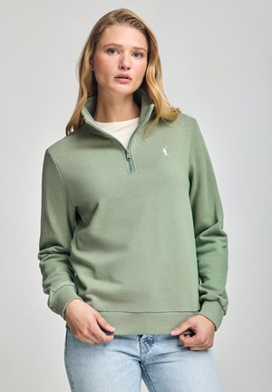 Sweat-shirt zippé vert clair avec un col montant, texture douce et logo blanc sur la poitrine gauche. Porté avec un jean bleu clair.