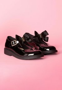 Pantofi din piele patent neagră cu vârfuri rotunde, având funde late din satin și catarame ajustabile pe vârf. Tălpi groase din cauciuc.