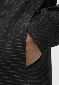 Juodas marškinys su tekstūruotu audiniu, kuriame yra mažų baltų trikampių raštų. Įeina šoninė kišenė ir mygtuko detalė.