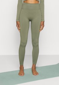 Olivgröna leggings med en hög ribbad midja, slät textur och en figurnära design, barfota på en ljusblå matta.