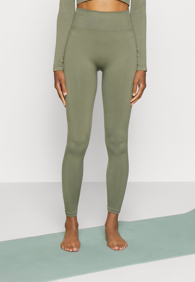 Olivgröna leggings med en hög ribbad midja, slät textur och en figurnära design, barfota på en ljusblå matta.