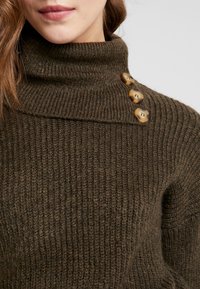 Gros plan sur une femme portant un pull à col roulé en maille côtelée marron avec trois boutons décoratifs marron sur l'épaule.