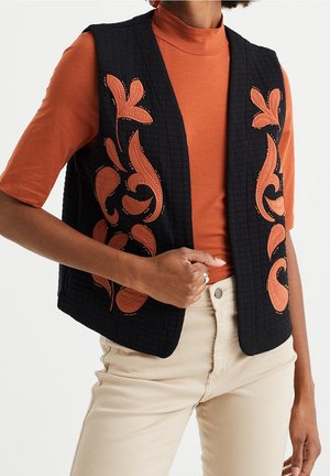 Vrouw draagt een zwart doorgestikt vest met oranje bloemenborduursel over een oranje top met korte mouwen en beige broek, hand rust op de taille.
