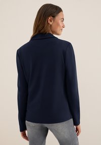 Mörkblå långärmad blazer med slagkrage, slät yta och smal passform, å paired with ljusgrå jeans. Minimalistisk design, inga synliga mönster.