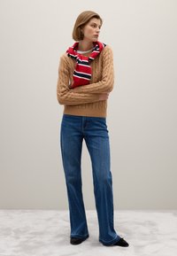 Suéter marrón de punto trenzado sobre una blusa beige, combinado con jeans acampanados azules y una bufanda a rayas rojas y navy atada al cuello.