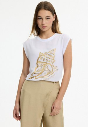 Joven mujer con camiseta blanca sin mangas con diseño de concha dorada y pantalones beige de talle alto sobre un fondo liso.
