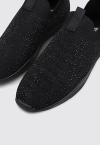 Zapatos negros tipo slip-on hechos de un material de malla transpirable con un patrón texturizado. Cuentan con aberturas elásticas y un diseño de punta redonda.