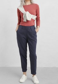 Rostfärgad långärmad topp kombinerad med marinblå tapered byxor. En vit tröja knuten avslappnat runt axlarna. Vita sneakers fulländar outfiten.