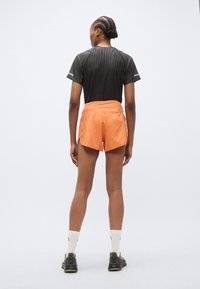 Chemise de sport noire avec des rayures, associée à un short orange avec des fentes latérales. Portée avec des chaussettes blanches et des chaussures sombres, vue de dos.