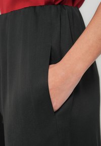Czarne spodnie z tkaniny o gładkiej fakturze, z boczną kieszenią i marszczonym pasem. Czerwony element górnej części jest częściowo widoczny u góry.