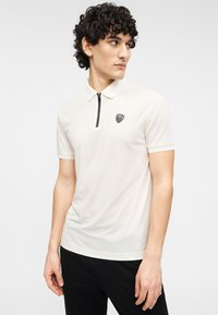 Φωτεινό μπεζ πόλο πουκάμισο με κοντά μανίκια, γιακά με φερμουάρ και μικρό μαύρο λογότυπο στο στήθος. Ομαλή υφή, σχεδιασμός στενής γραμμής.