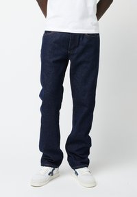Donkerblauwe denim jeans met een recht model, voorzien van subtiele stiksels en standaard voorzakken. Draagt met witte sneakers.