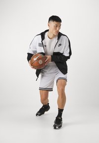 Jugador de baloncesto de Nike vistiendo un chándal negro y blanco, sosteniendo un balón de baloncesto naranja. El atuendo presenta una marca pronunciada y franjas en contraste.