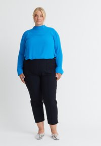 Blaue Bluse mit langen Ärmeln und hohem Kragen, kombiniert mit schwarzen, schmal zulaufenden Hosen, abgerundet mit silbernen Ballerinas. Glattes Material; elegantes Design.