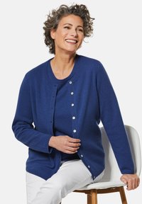 Blauer Cardigan aus weichem Stoff mit rundem Halsausschnitt und Knopfverschluss. Hat lange Ärmel und eine lockere Passform, kombiniert mit einem blauen Oberteil.