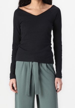 Top lungo a maniche nere a coste con design a V, abbinato a pantaloni verdi drappeggiati con dettaglio di legatura frontale.