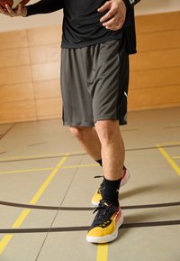 Pantalones cortos deportivos grises con un logo negro, combinados con zapatos amarillos y negros. El jugador sostiene una pelota de baloncesto, de pie en una cancha de gimnasio con marcas.